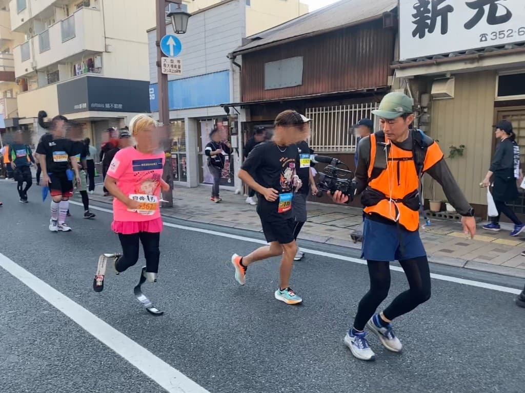 都市のマラソンコースで義足ランナーを撮影するハンドヘルドカメラのカメラマン
